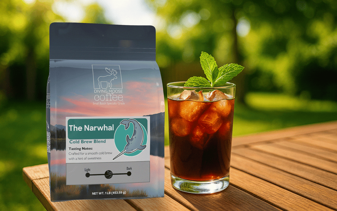 Bag of Diving Moose Coffee's The Narwhal Cold Brew Blend next to a glass of iced cold brew coffee garnished with mint on a wooden table outdoors